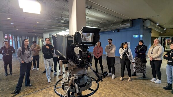 Eine Gruppe von Menschen steht in einem großen, hellen Raum mit industriellem Design. Im Vordergrund ist eine professionelle Fernsehkamera auf einem Stativ zu sehen, die auf die Gruppe gerichtet ist. Die Personen sind unterschiedlich gekleidet und stehen in einer lockeren Formation. Einige blicken in Richtung Kamera, andere unterhalten sich. Der Raum hat eine hohe Decke mit sichtbaren Rohren und Beleuchtungselementen.