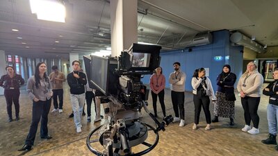 Eine Gruppe von Menschen steht in einem großen, hellen Raum mit industriellem Design. Im Vordergrund ist eine professionelle Fernsehkamera auf einem Stativ zu sehen, die auf die Gruppe gerichtet ist. Die Personen sind unterschiedlich gekleidet und stehen in einer lockeren Formation. Einige blicken in Richtung Kamera, andere unterhalten sich. Der Raum hat eine hohe Decke mit sichtbaren Rohren und Beleuchtungselementen.