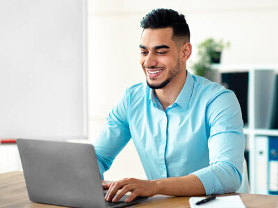 Ein lächelnder junger Mann mit einem hellblauen Hemd sitzt an einem Schreibtisch und arbeitet an einem Laptop. Der Hintergrund zeigt ein Büro mit einem Regal und einer Pflanze.