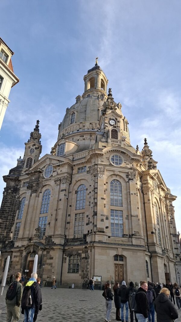 Die Dresdner Frauenkirche aus der Froschperspektive, mit großer Kuppel und barocker Fassade, davor Menschen auf dem Platz.