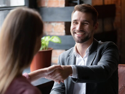 Ein Mann mit Bart und einem grauen Anzug sitzt lächelnd an einem Tisch und schüttelt einer Frau die Hand. Die Frau, deren Rücken zu sehen ist, hat lange blonde Haare und trägt ein burgunderfarbenes Oberteil. Beide scheinen sich in einem Büro oder Besprechungsraum zu befinden, der mit Pflanzen und modernen Möbeln ausgestattet ist.