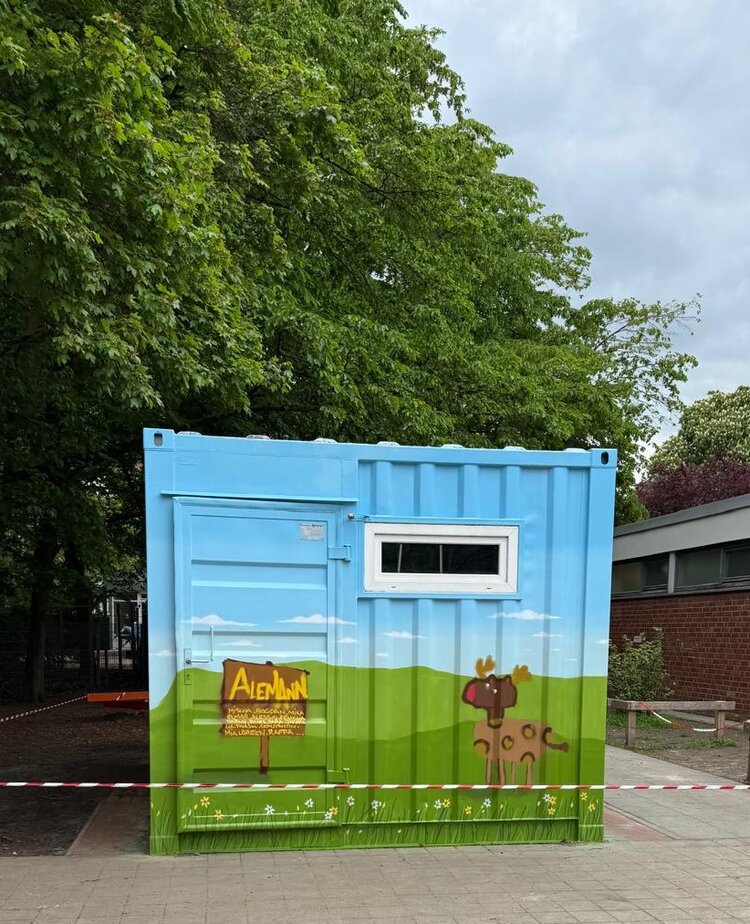 Ein bemalter Container, der als Geräteschuppen dient. Er ist mit einer grünen Landschaft, einem cartoonhaften Reh und einem Holzschild mit der Aufschrift „Alemann“ bemalt ist. Im Hintergrund sind Bäume und ein bewölkter Himmel zu sehen. Der Container steht auf einem gepflasterten Platz.