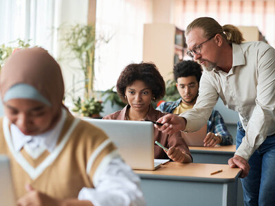 In einem Klassenzimmer oder Schulungsraum unterstützt ein älterer Mann mit Brille und Zopf zwei junge Erwachsene bei der Arbeit an ihren Laptops. Der Mann zeigt mit einem Stift auf den Bildschirm eines Laptops, während eine junge Frau mit lockigem Haar konzentriert zuschaut. Ein weiterer junger Mann im Hintergrund arbeitet ebenfalls an einem Laptop. Links im Bild ist eine Person mit Kopfbedeckung zu sehen, die ebenfalls an einem Laptop arbeitet. Der Raum ist hell und freundlich, mit Pflanzen im Hintergrund. Die Szene vermittelt eine Lern- oder Arbeitsatmosphäre, in der Unterstützung und Zusammenarbeit im Vordergrund stehen.