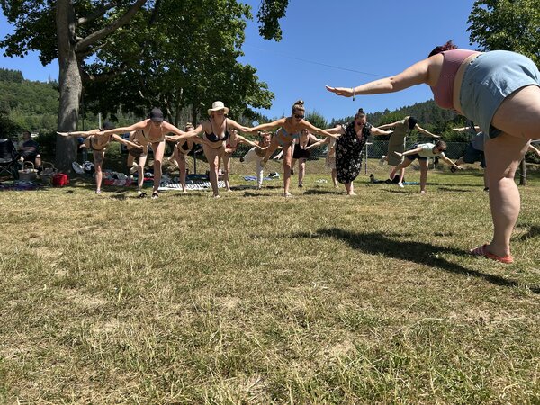 Auf einer Wiese praktiziert eine Gruppe junger Menschen Yoga: sie alle stehen auf einem Bein und haben die Arme wie zum Fliegen ausgebreitet.