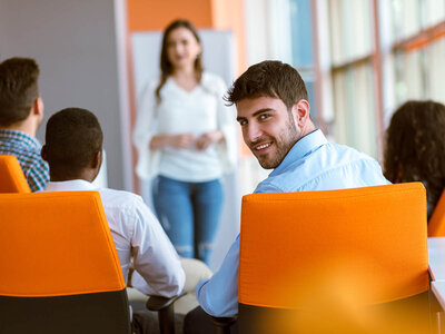 Eine Gruppe von Menschen sitzt in orangefarbenen Stühlen und blickt zu einer Person, die vorne im Raum steht. Die Person scheint eine Präsentation zu halten oder zu der Gruppe zu sprechen. Im Hintergrund sind große Fenster und eine orangefarbene Wand zu sehen.