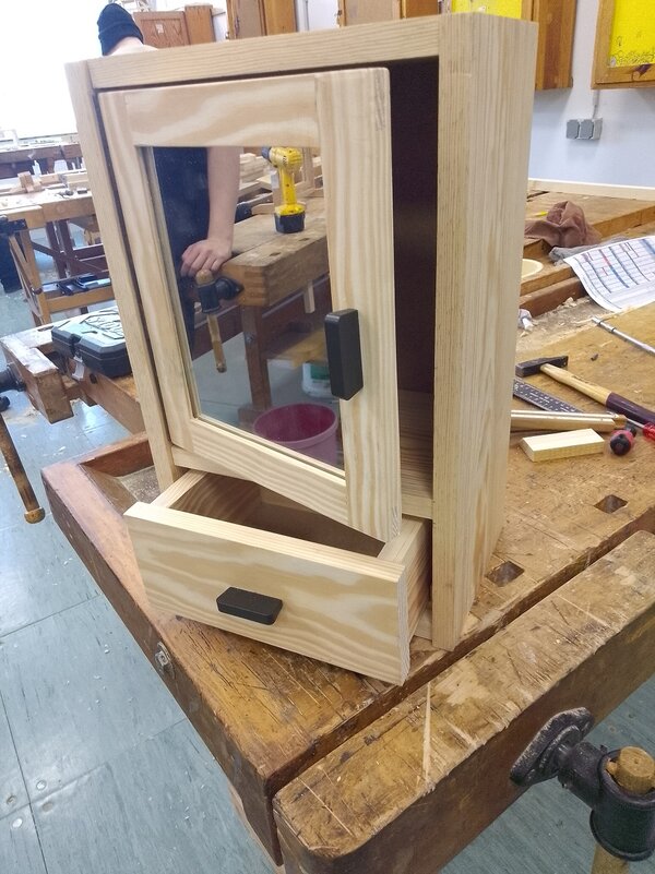 Kleines Holzschränkchen auf einer Werkbank mit Glastür und schwarzem Griff; darunter ist eine Schublade leicht geöffnet, Werkzeuge liegen im Hintergrund.