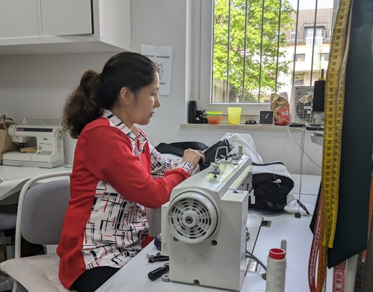 Eine Frau mit Zopf näht mit einer Nähmaschine ein Kleidungsstück. Rechts im Vordergrund hängen Maßbänder an der Wand. Im Hintergrund ist links ein Tisch mit einer weiteren Nähmaschine, rechts ist ein Fenster zu einem Hof mit Bäumen.