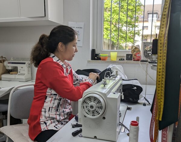 Eine Frau mit Zopf näht mit einer Nähmaschine ein Kleidungsstück. Rechts im Vordergrund hängen Maßbänder an der Wand. Im Hintergrund ist links ein Tisch mit einer weiteren Nähmaschine, rechts ist ein Fenster zu einem Hof mit Bäumen.