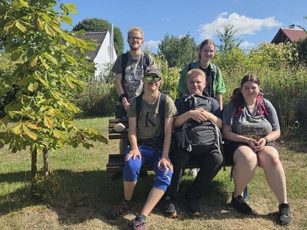 Fünf Personen sitzen auf einer Holzbank in einem grünen Garten. Im Hintergrund sind Bäume und ein kleines Haus mit rotem Dach zu erkennen. Die Stimmung wirkt entspannt und sommerlich.