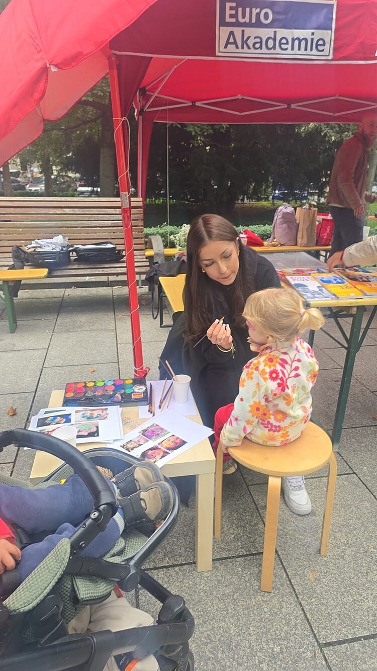 Eine Mitarbeiterin schminkt am Kinderschminkstand der Euro-Schulen ein kleines Mädchen, das auf einem Hocker sitzt und nur noch hinten zu sehen ist.