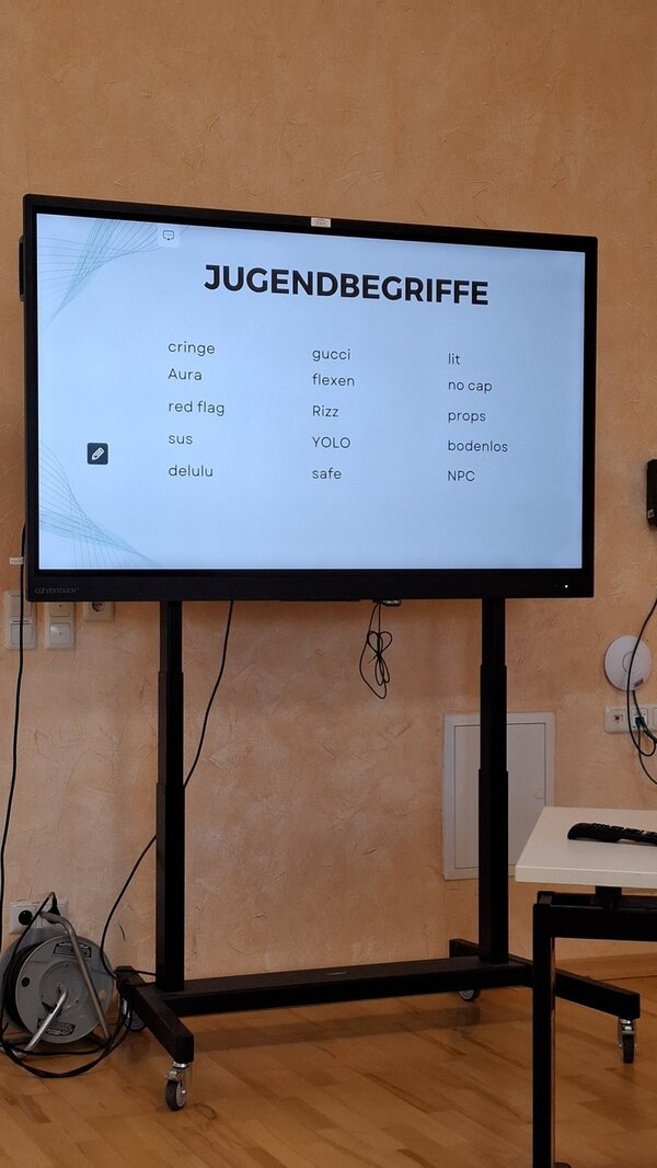 Ein großer, auf einem schwarzen fahrbaren Ständer montierter Bildschirm steht vor einer orangefarbenen Wand in einem Schulungsraum. Auf dem Bildschirm ist eine Präsentationsfolie mit dem Titel 'JUGENDBEGRIFFE' zu sehen, darunter sind verschiedene Begriffe in drei Spalten aufgelistet: 'cringe', 'Aura', 'red flag', 'sus', 'delulu', 'gucci', 'flexen', 'Rizz', 'YOLO', 'safe', 'lit', 'no cap', 'props', 'bodenlos', 'NPC'. Am Boden neben dem Bildschirm liegen Kabel und eine graue Tasche. Rechts neben dem Bildschirm steht ein weißer Tisch mit einem schwarzen Gegenstand darauf. Die Szene zeigt eine Inhouse-Weiterbildung für Mitarbeitende zum Thema 'Kommunikation mit der Generation Z' an den Euro-Schulen Hoyerswerda.