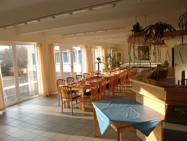 Heller Speisesaal mit großen Fenstern, langen eingedeckten Holztischen und Stühlen, dekoriert mit Pflanzen und Luftschlangen.