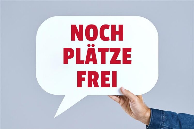 Eine Hand hält ein weißes Sprechblasen-Schild mit der Aufschrift „NOCH PLÄTZE FREI“ in roten Großbuchstaben.