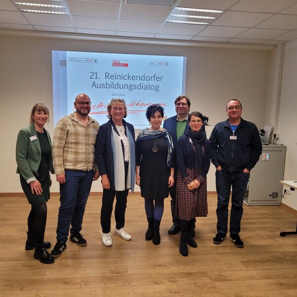 Eine Gruppe von sieben Personen steht im Seminarraum vor einer großen Projektionsfläche, auf der ‚21. Reinickendorfer Ausbildungsdialog‘ und das Datum angezeigt werden. Die Gruppe lächelt in die Kamera und steht locker nebeneinander. Der Raum hat einen hellen Boden, neutrale Wände und eine moderne Präsentationstechnik. Das Bild dokumentiert den gemeinsamen Austausch beim 21. Reinickendorfer Ausbildungsdialog an den Euro-Schulen Berlin.