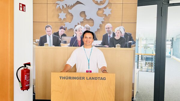 Zu sehen ist der Plenarsaal das Landtags in Thüringen. In der Mitte ist das Rednerpult, an dem ein Mann in weißem Hemd steht und eine Rede hält. Dahinter sitzen die Landtagspräsidentin und ihre Unterstützer.