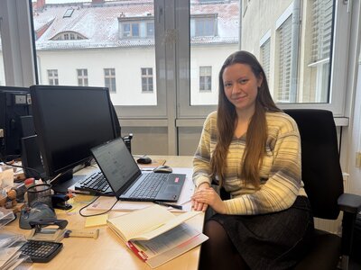 Eine junge Frau mit hellem Oberteil sitzt an einem Schreibtisch mit einem Notebook und schaut leichtnach links gedreht in die Kamera.