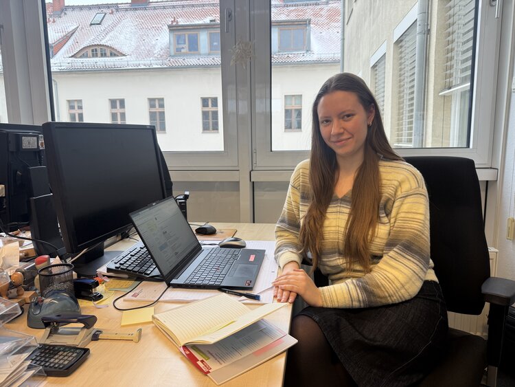 Eine junge Frau mit hellem Oberteil sitzt an einem Schreibtisch mit einem Notebook und schaut leichtnach links gedreht in die Kamera.