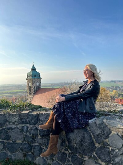 Eine lachende junge Frau mit blondem Haar und Kleid sitzt auf einer erhöhten Mauer. Im Hintergrund ist ein Kirchturm vor einem blauen Himmel und der weiten Landschaft zu sehen.