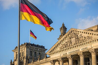 Einbürgerungstests ab Juli auch an den Euro-Schulen Berlin möglich Am Deutschen Reichstag wehen zwei Deutschlandflaggen, darüber strahlt ein fast wolkenloser Himmel.