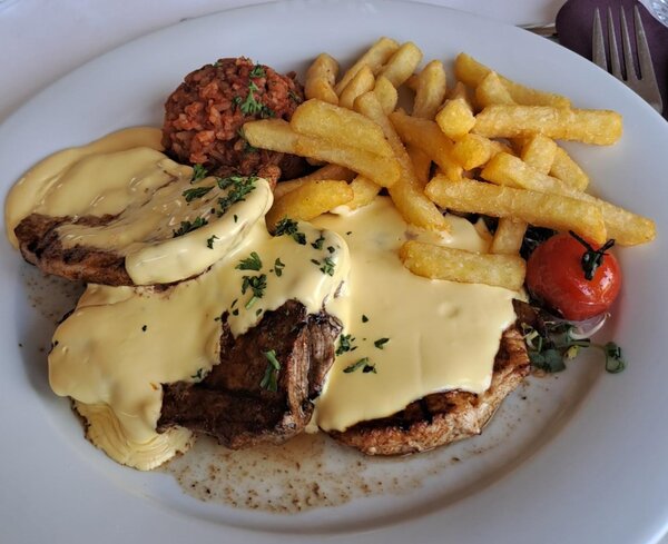 Ein Teller mit einem Hauptgericht: Ein Stück Fleisch mit Käse überbacken, dazu Pommes frites, ein kleiner Berg Reis und eine Kirschtomate als Garnitur.