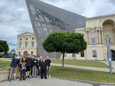 Die Teilnehmenden stehen vor dem beeindruckenden Gebäude des Militärhistorischen Museums in Dresden, das in der architektonischen Stilrichtung des Dekonstruktivismus gebaut wurde.