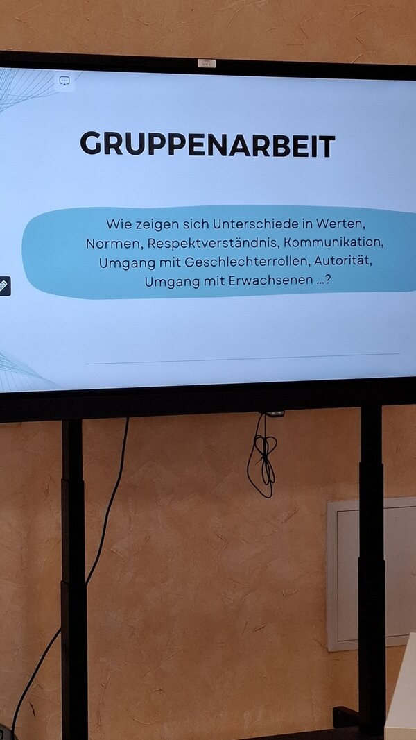 Ein großer Bildschirm zeigt eine Präsentationsfolie mit dem Titel 'GRUPPENARBEIT'. Darunter steht in einem hellblauen, abgerundeten Rechteck der Text: 'Wie zeigen sich Unterschiede in Werten, Normen, Respektverständnis, Kommunikation, Umgang mit Geschlechterrollen, Autorität, Umgang mit Erwachsenen ...?'. Der Bildschirm steht vor einer hellbraunen Wand, und unter dem Bildschirm sind zwei Kabel sichtbar, die nach unten hängen.