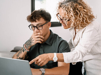 Ein junger Mann mit Brille, Tattoos auf dem Arm und einem dunklen Polo-Shirt sitzt vor einem Laptop. Er stützt sein Kinn auf die Hand und wirkt nachdenklich. Eine Frau mit lockigen Haaren, Brille und einem weißen Hemd steht neben ihm und zeigt auf den Bildschirm des Laptops. Beide scheinen konzentriert an einer Aufgabe zu arbeiten oder etwas zu besprechen. Der Hintergrund ist neutral und zeigt einen Innenraum, möglicherweise ein Büro.