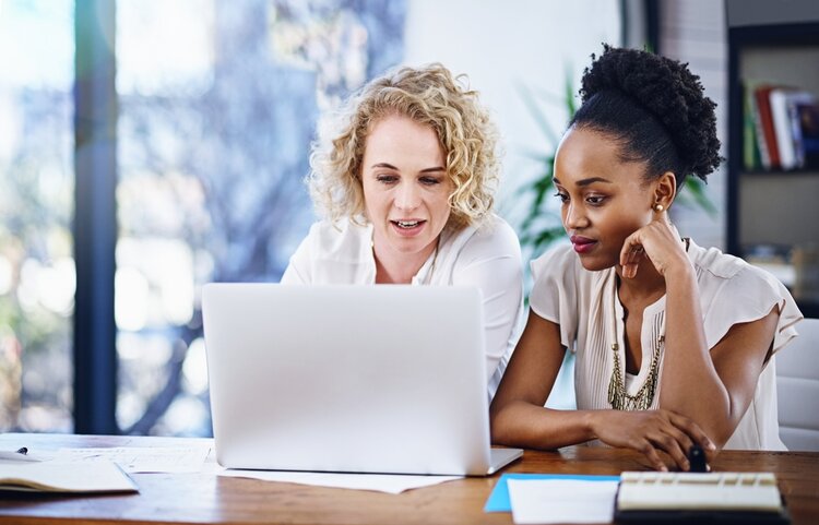 Zwei junge Frauen sitzen vor einem Notebook und überlegen etwas.