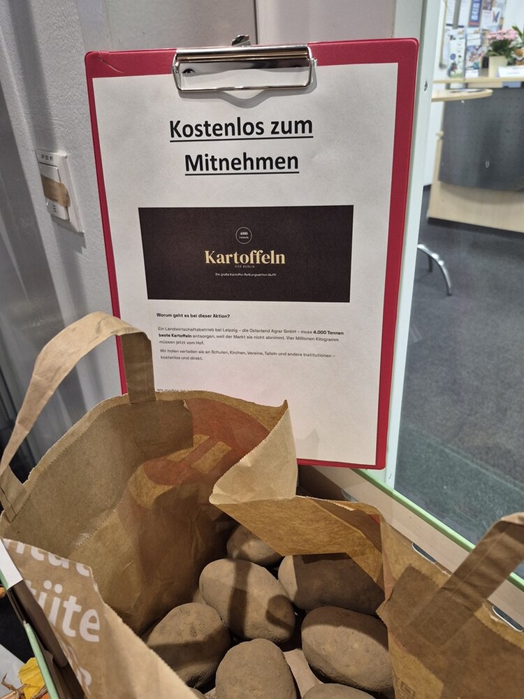 Eine braune Papiertüte mit mehreren Kartoffeln steht auf einem Tisch. Dahinter hängt ein Klemmbrett mit einem weißen Blatt Papier, auf dem in großer, fettgedruckter Schrift steht: 'Kostenlos zum Mitnehmen'. Darunter befindet sich ein schwarzer Kasten mit dem Wort 'Kartoffeln' in weißer Schrift. Im Hintergrund sind Teile eines Büros mit Schreibtisch, Computer und weiteren Gegenständen zu sehen.