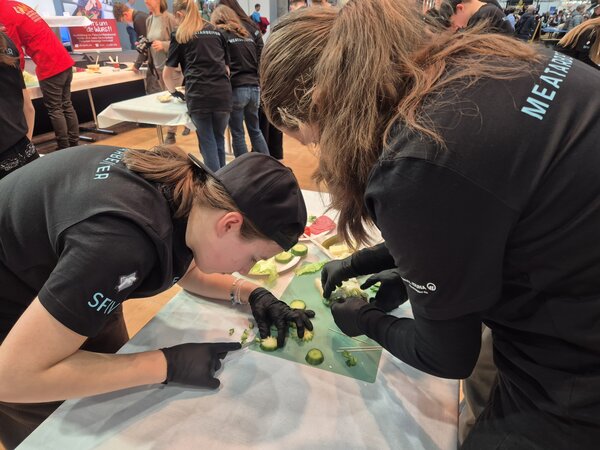 Zwei Schülerinnen mit schwarzen Gummihandschuhen sind über ein Schneidebrett gebeugt und schneiden Gurkenscheiben und Salat.