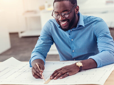 Ein lächelnder Mann mit Brille und blauem Hemd sitzt an einem Tisch und arbeitet konzentriert an einem großen Bauplan oder einer technischen Zeichnung. Er hält einen Bleistift in der Hand und trägt eine Armbanduhr am linken Handgelenk. Der Hintergrund zeigt einen hellen, modernen Raum, möglicherweise ein Büro oder ein Planungsbüro.