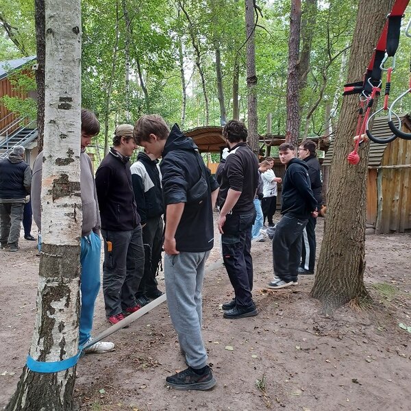 Eine Gruppe von Auszubildenden steht entlang eines Bandes, das zwischen zwei Baumstämmen gespannt ist. Im Hintergrund sind weitere Bäume und Holzgebäude zu sehen. Vorne rechts hängen Klettergriffe an einem Baum.