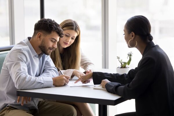 Drei Personen sitzen an einem Tisch in einem hellen, modernen Büro. Eine Beraterin zeigt auf ein Dokument, während ein junger Mann dieses unterzeichnet. Neben ihm sitzt eine junge Frau, die dem Gespräch aufmerksam folgt. Auf dem Tisch steht ein kleiner Kaktus in einem weißen Blumentopf.
