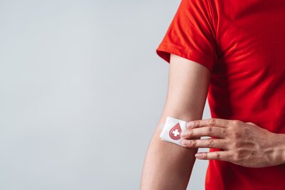 Ein Mann in rotem T-Shirt hälte sich an die rechte Armbeuge ein Pflaster mit einem Blutstropfen drauf.