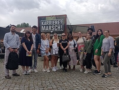 Besuch der JOBinale Die heterogene Gruppe der Teilnehmenden des B2-Berufssprachkurses an den Euro-Schulen Potsdam posieren gemeinsam und gut gelaunt vor der JOBinale-Messe für ein Gruppenfoto.