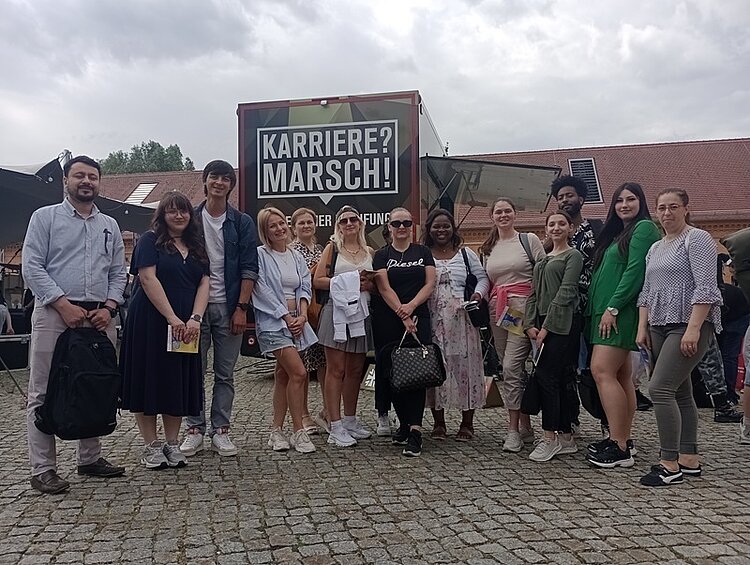 Besuch der JOBinale Die heterogene Gruppe der Teilnehmenden des B2-Berufssprachkurses an den Euro-Schulen Potsdam posieren gemeinsam und gut gelaunt vor der JOBinale-Messe für ein Gruppenfoto.
