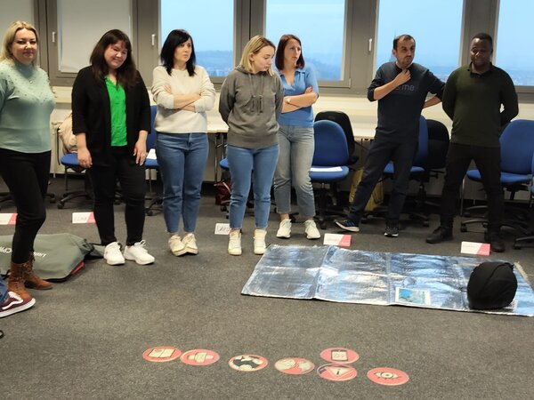 Eine Gruppe von sieben Personen steht in einem Klassenzimmer und beobachtet eine Übungssituation zur Ersten Hilfe. Auf dem Boden liegt eine Rettungsdecke mit Symbolen für verschiedene Notfallmaßnahmen. Die Teilnehmer wirken konzentriert und aufmerksam.