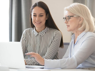 Zwei Frauen sitzen nebeneinander und arbeiten gemeinsam an einem Laptop. Eine Frau trägt ein gestreiftes Hemd und die andere ein blaues Hemd mit einer Brille. Beide konzentrieren sich auf den Bildschirm, während sie etwas auf dem Laptop betrachten oder bearbeiten.