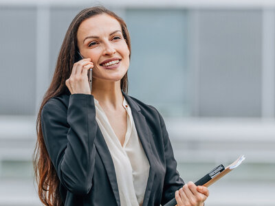 Eine lächelnde Person mit langen, dunklen Haaren und einer dunklen Blazerjacke über einem weißen Oberteil telefoniert mit einem Smartphone und hält dabei einen Bündel mit Dokumenten und ein Notebook in der Hand. Im Hintergrund ist ein unscharfes Büroumfeld zu erkennen.
