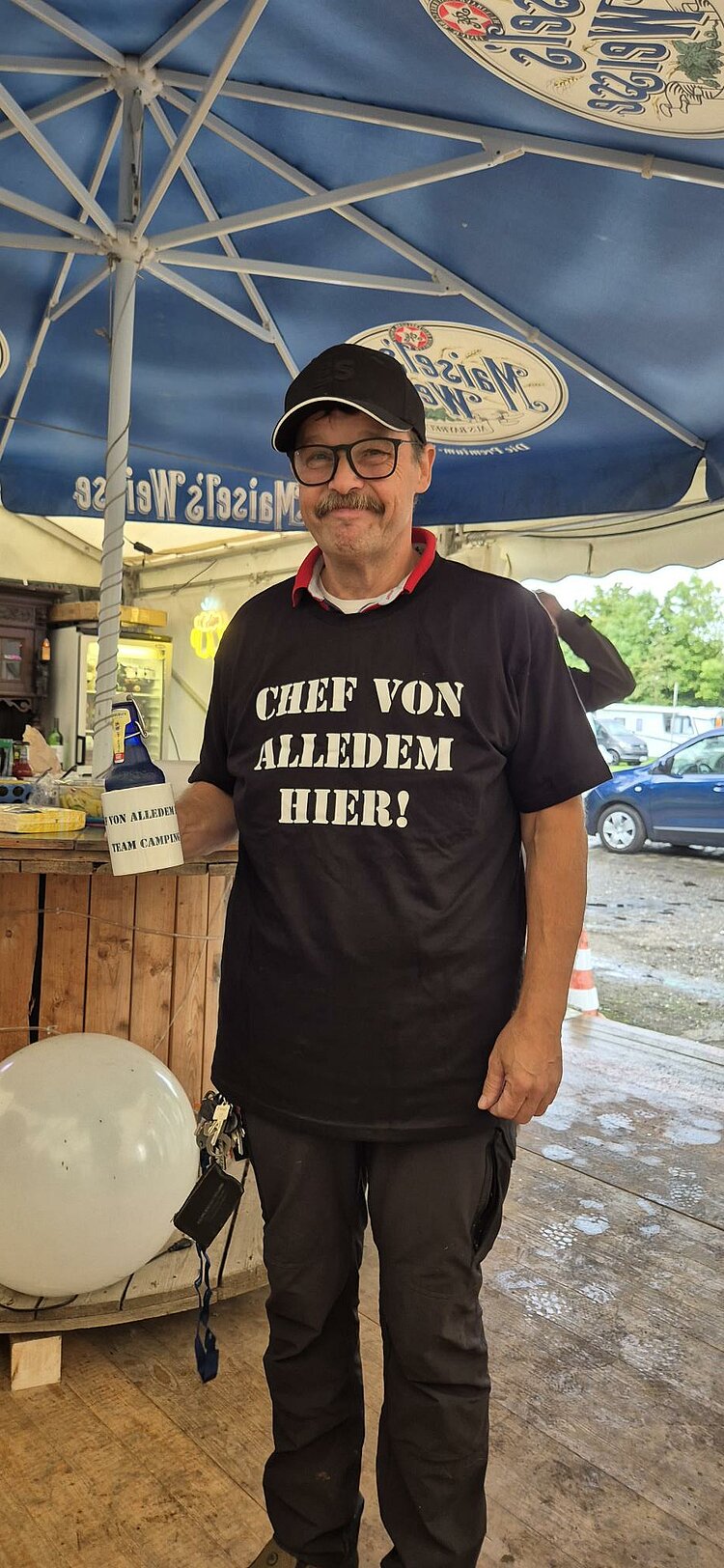 Ein Mann mit Brille und Baseballkäppi grinst verlegen in die Kamera. Er ist bekleidet mit einem T-Shirt mit der Aufschrift "Chef von alledem hier!" und hat eine Tasse mit dem zusätzlichen Text "Team Camping", in der eine Henkelbierflasche steckt.