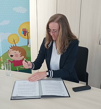 Unterzeichnung des Kooperationsvertrages im Projekt „Resilienz in der Kinder- und Jugendarbeit (Kita-Resilienz)“ Frau Anke Mädler sitzt an einem Tisch, unterschreibt ein Dokument in einer Mappe. Neben ihr steht ein Glas Wasser, im Hintergrund ein buntes Plakat mit einer Kinderzeichnung.