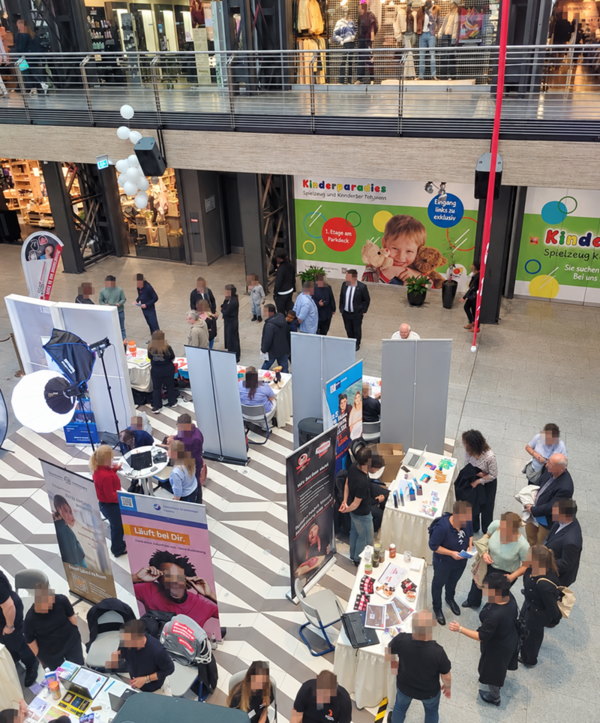 Messebereich in einer Einkaufspassage mit mehreren Ständen, an denen Menschen im Gespräch sind. Die Stände sind mit Roll-ups, Flyern und Werbematerialien ausgestattet. Im Hintergrund befinden sich große Werbeplakate mit bunten Motiven und dem Schriftzug 'Kinderparadies'. Die Szenerie ist von oben aufgenommen, sodass die Anordnung der Stände und die Bewegungen der Besucher gut erkennbar sind. Die Umgebung wirkt modern und offen, mit einer Galerie im oberen Bereich und Schaufenstern im Hintergrund.