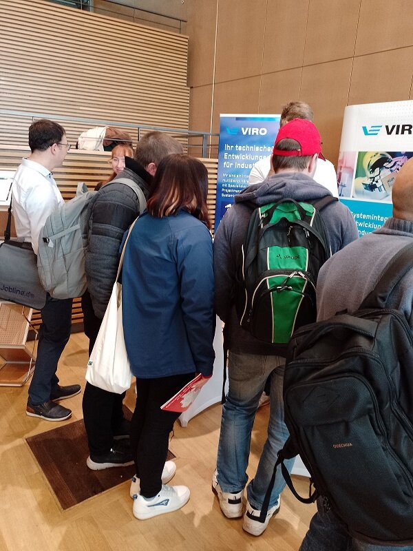 Kleingruppe Messebesucher*innen steht dicht vor dem VIRO-Stand und liest die Infos auf zwei blauen Roll-ups.