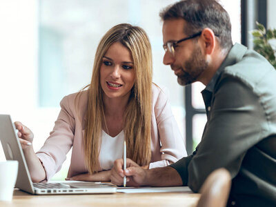 Eine Frau und ein Mann sitzen an einem Tisch in einem Büro oder Besprechungsraum. Die Frau, mit langen blonden Haaren und einem hellen Blazer, hält einen Stift in der Hand und zeigt ihrem Kollegen etwas auf einem Laptop-Bildschirm. Der Mann, mit Brille und einem dunklen Hemd, beobachtet aufmerksam den Bildschirm und hört ihr zu. Beide wirken konzentriert und engagiert. Auf dem Tisch liegen ein Notizblock und ein Kaffeebecher. Der Raum wirkt modern und hell, mit großen Fenstern im Hintergrund. Die Atmosphäre ist professionell und kooperativ.