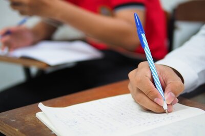 Nahaufnahme eines Schülers, der eine Prüfung ablegt und mit einem blau gestreiften Stift in ein Notizbuch schreibt. Der Fokus liegt auf den Themen Bildung, Prüfungen und Lernumgebungen im Klassenzimmer.