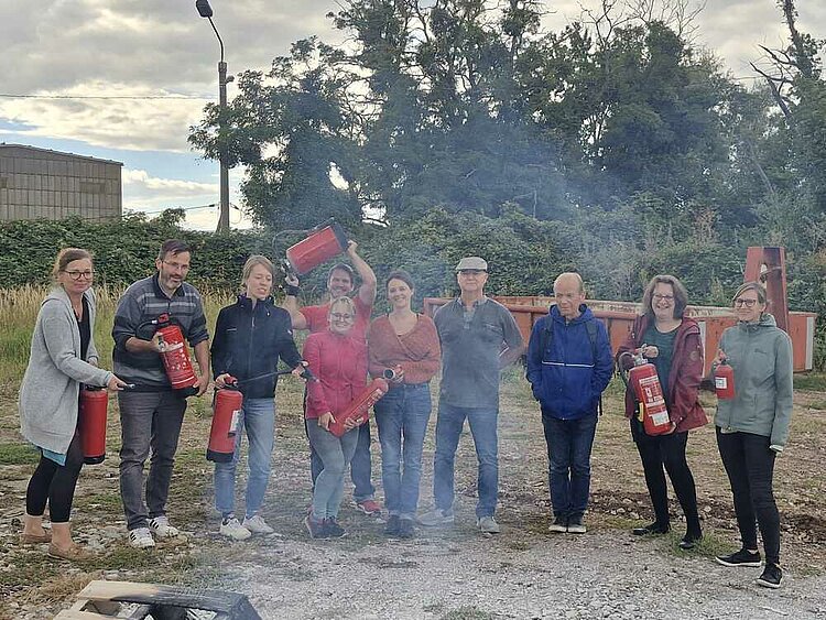 Mit Wasser gegen Feuer? Auf einem Schotterplatz stehen zehn Männer und Frauen, von denen fast all unterschiedliche Feuerlöscher in der Hand haben. Diese sind auf ein Feuer vor ihnen gerichtet, das nicht im Bild ist - allerdings kann man es durch eine angekokelte Palette und Rauch im Vordergrund erahnen.