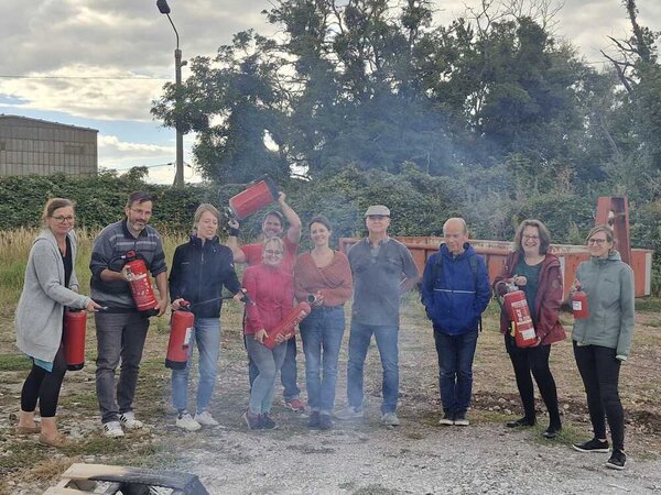 Auf einem Schotterplatz stehen zehn Männer und Frauen, von denen fast all unterschiedliche Feuerlöscher in der Hand haben. Diese sind auf ein Feuer vor ihnen gerichtet, das nicht im Bild ist - allerdings kann man es durch eine angekokelte Palette und Rauch im Vordergrund erahnen.