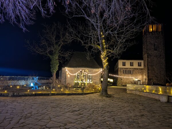 Nächtlicher Burghof mit Kopfsteinpflaster, Bäumen und warmen Lichterketten, kleine historische Gebäude und Turm im Hintergrund.