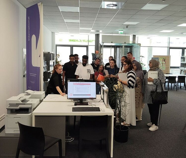 Gruppe IK-359 zu Besuch im BIZ Eine Gruppe von Teilnehmenden des Integrationskurses steht um einen Computerarbeitsplatz im Berufsinformationszentrum und erhält Anleitungen von einer Beraterin.