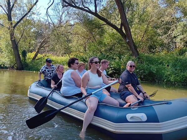 Eine Gruppe von sieben Männern und Frauen sitzt in und auf dem Rand eines blauen Schlauchboots. Einige Personen haben Sonnenbrillen und Kopfbedeckungen auf, sie halten Ruder in der Hand. Durch die Bäume am Ufer liegt das Boot im Schatten.